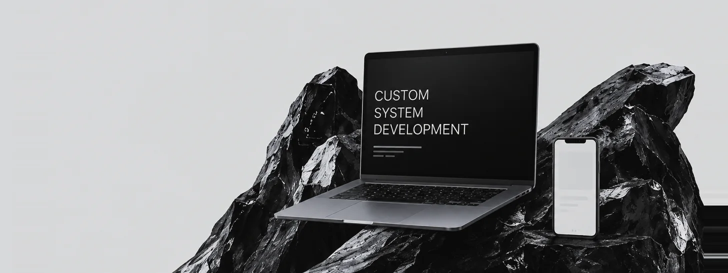 黒い岩の上に置かれたノートPCとスマートフォン。画面に「CUSTOM SYSTEM DEVELOPMENT」の文字
